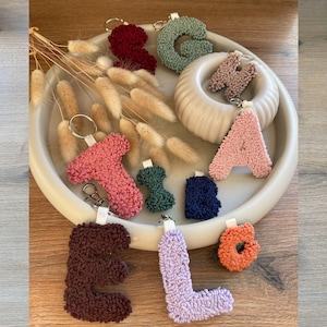 Könnte beinhalten: Eine Auswahl an strukturierten Buchstaben-Schlüsselanhängern in Farben wie Rosa, Grün und Blau. Die Schlüsselanhänger liegen auf einem weißen Tablett mit Trockenblumen. Ideal zum Personalisieren von Taschen oder Schlüsseln.