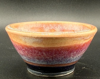 Puede incluir: Un cuenco pequeño de cerámica con un esmalte multicolor que presenta bandas horizontales en tonos beige, rojo oscuro, morado y azul marino. El borde interior muestra un tono beige más claro y la superficie exterior es lisa y brillante.