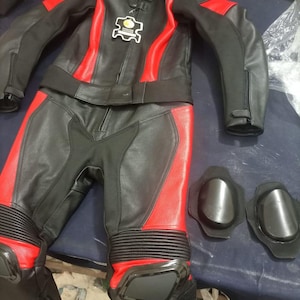 May include: Black and red motorcycle racing suit with matching knee pads. The suit features a black leather jacket with red accents and matching pants. The jacket has a small patch with text. The knee pads are black.