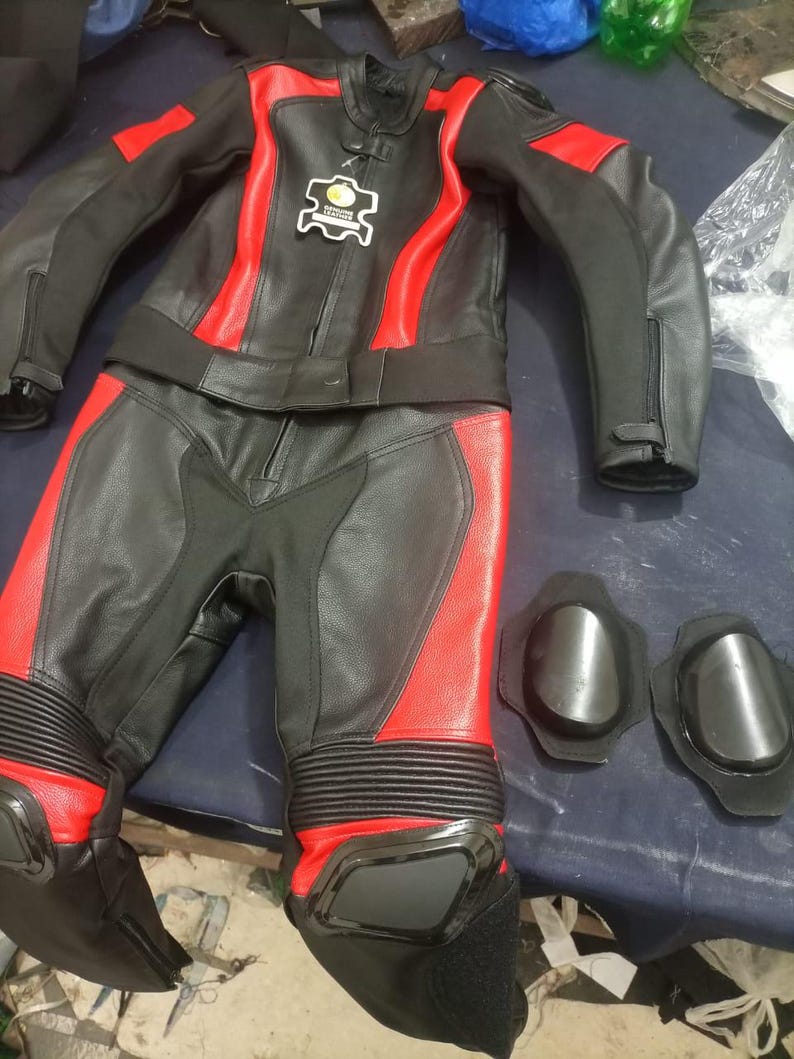 May include: Black and red motorcycle racing suit with matching knee pads. The suit features a black leather jacket with red accents and matching pants. The jacket has a small patch with text. The knee pads are black.