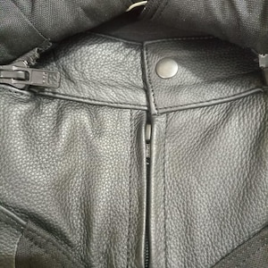 May include: Close-up of black leather motorcycle pants with a zipper and snap closure. The leather has a textured appearance, and the pants have black fabric accents. The zipper is silver, and the snap is a dull silver color.