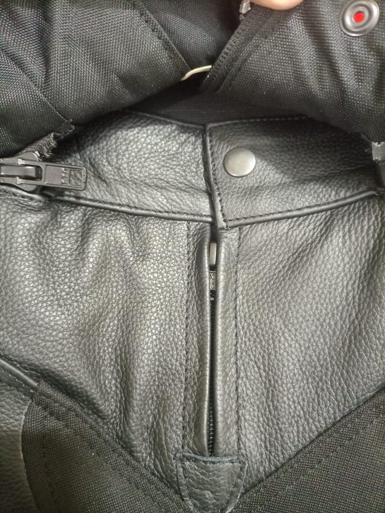 May include: Close-up of black leather motorcycle pants with a zipper and snap closure. The leather has a textured appearance, and the pants have black fabric accents. The zipper is silver, and the snap is a dull silver color.