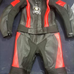 May include: Black and red leather motorcycle suit. The suit includes a jacket and pants. The jacket has red accents on the shoulders and sides. The pants have red accents on the lower legs. The suit has a tag that says "Genuine Leather".