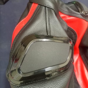May include: Close-up of a black and red leather motorcycle jacket. The jacket features a black padded elbow protector with a shiny black border. A red stripe runs down the sleeve.