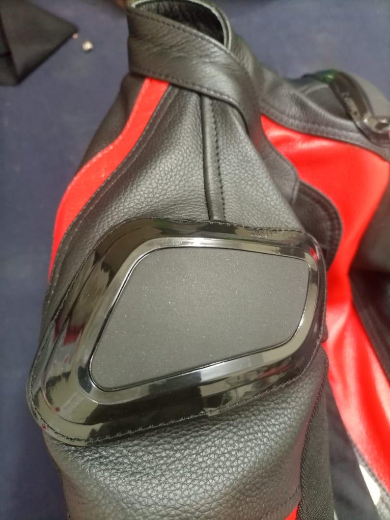 May include: Close-up of a black and red leather motorcycle jacket. The jacket features a black padded elbow protector with a shiny black border. A red stripe runs down the sleeve.