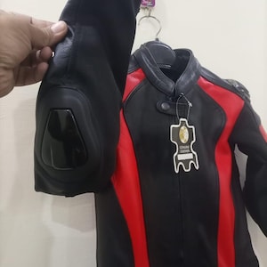 May include: Black and red leather motorcycle jacket with a stand-up collar. The jacket has red panels on the shoulders and sides. The jacket is hanging on a hanger. The sleeve is being held up by a hand.
