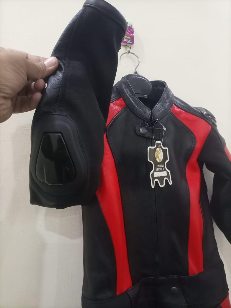 May include: Black and red leather motorcycle jacket with a stand-up collar. The jacket has red panels on the shoulders and sides. The jacket is hanging on a hanger. The sleeve is being held up by a hand.