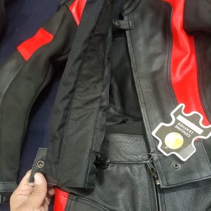 May include: Black and red leather motorcycle jacket and pants set. The jacket has red accents on the shoulders and sleeves. The pants have red accents on the sides. A tag that says "GENUINE LEATHER" is attached to the jacket.
