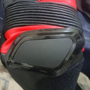 May include: Close-up of a black and red motorcycle boot. The boot features a black protective panel, ribbed detailing, and a red leather upper. The design suggests a focus on protection and performance.