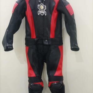 May include: Black and red motorcycle racing suit. The suit features a black base with red accents on the shoulders, arms, and legs. It has a high collar, a zipper closure, and protective padding on the knees and shoulders. A tag with the words "Genuine Leather" is attached.