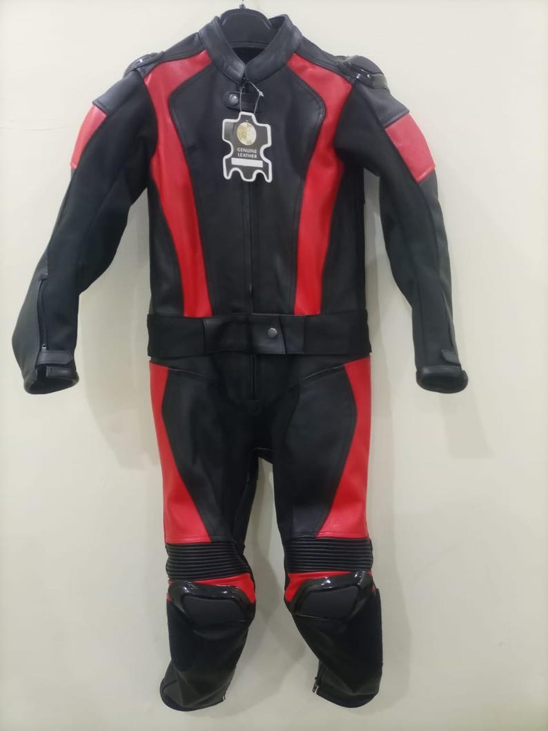 May include: Black and red motorcycle racing suit. The suit features a black base with red accents on the shoulders, arms, and legs. It has a high collar, a zipper closure, and protective padding on the knees and shoulders. A tag with the words "Genuine Leather" is attached.