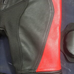 May include: Close-up of a black and red leather motorcycle pant leg. The design features a black main panel with a red side panel. The lower leg has ribbed detailing. A black knee protector is visible.