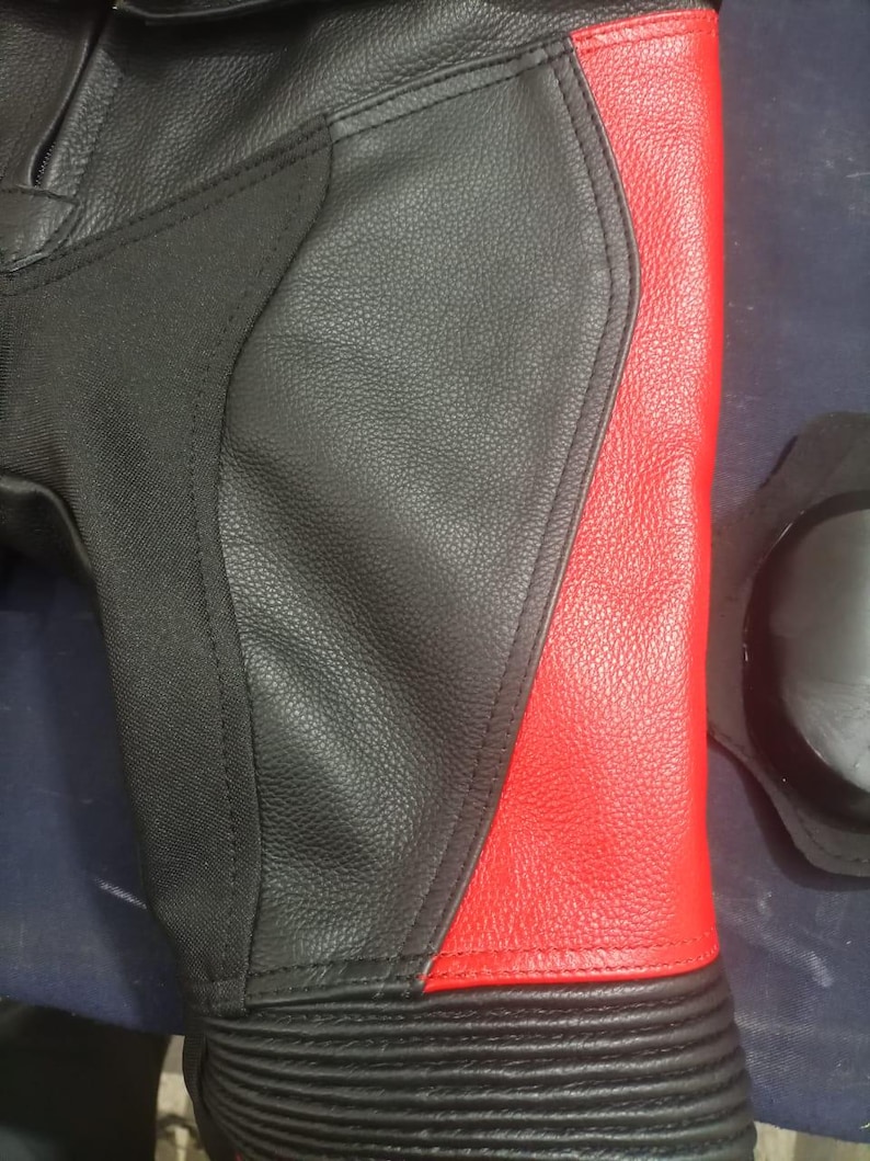 May include: Close-up of a black and red leather motorcycle pant leg. The design features a black main panel with a red side panel. The lower leg has ribbed detailing. A black knee protector is visible.