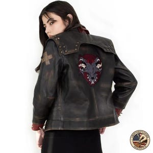 May include: Distressed black leather jacket with a large embroidered wolf patch on the back. The jacket features a unique collar design, studded details, and a red lining. A cross patch is on the left shoulder.