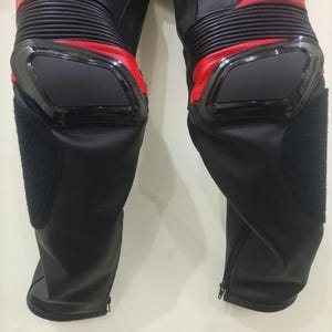 May include: Black and red motorcycle pants with protective knee pads. The pants have a zippered closure at the ankles and ribbed panels above the knees. The knee pads are black and outlined with a glossy black trim.