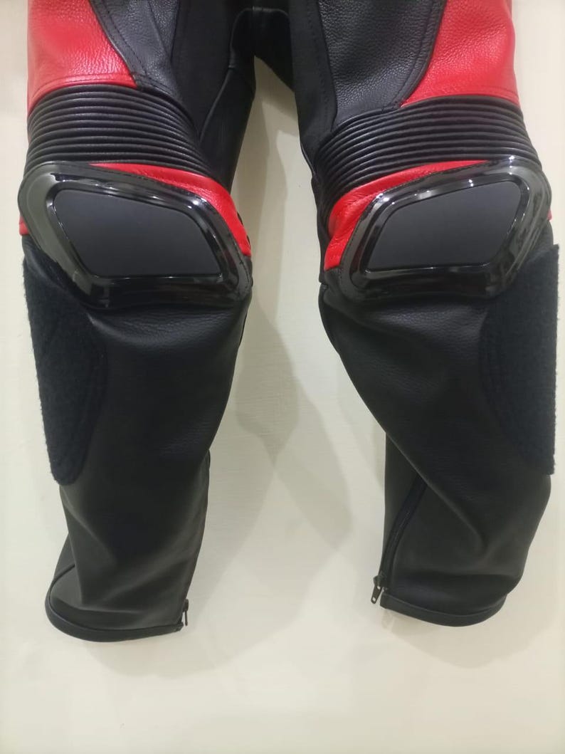 May include: Black and red motorcycle pants with protective knee pads. The pants have a zippered closure at the ankles and ribbed panels above the knees. The knee pads are black and outlined with a glossy black trim.