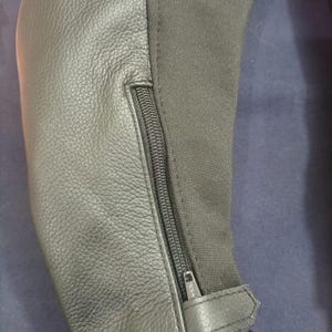 May include: Close-up of a black leather and fabric garment. The leather has a textured appearance, and a black zipper runs along the seam. A small leather strap with a buckle is visible at the bottom.