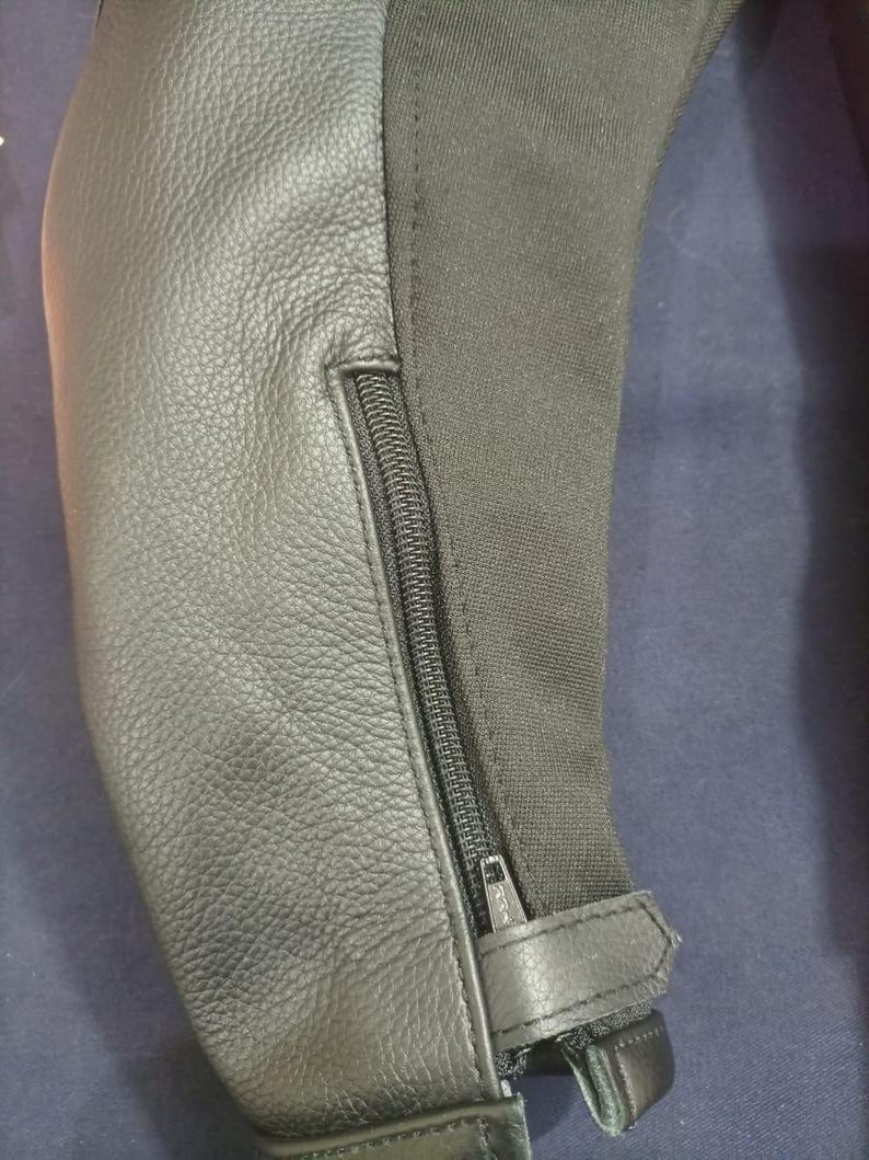 May include: Close-up of a black leather and fabric garment. The leather has a textured appearance, and a black zipper runs along the seam. A small leather strap with a buckle is visible at the bottom.