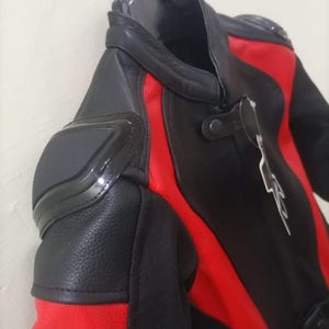 May include: Black and red motorcycle jacket with protective padding on the shoulders. The jacket features a leather exterior with red accents and a high collar. The jacket is hanging on a metal hanger.