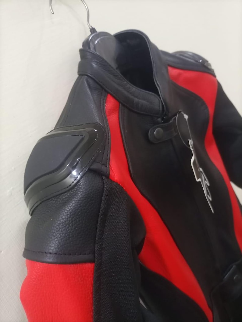 May include: Black and red motorcycle jacket with protective padding on the shoulders. The jacket features a leather exterior with red accents and a high collar. The jacket is hanging on a metal hanger.