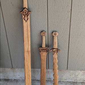 May include: Three wooden swords of varying sizes, each with intricate carved details. The largest sword is approximately 91 cm long, featuring a diamond-shaped pommel and a decorative crossguard. The smaller swords have circular and stylized designs.