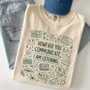 Op de afbeelding: Een crèmekleurig t-shirt met korte mouwen en donkergroene lijntekeningen van diverse communicatiemiddelen en de tekst "HOWEVER YOU COMMUNICATE, I AM LISTENING." Het ontwerp bevat iconen van een toetsenbord, tekstballonnen en ogen.
