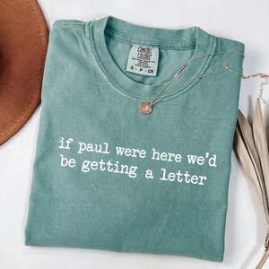 Może przedstawiać: Miętowa koszulka z białym napisem "if paul were here we'd be getting a letter". Złoty naszyjnik z wisiorkiem spoczywa na koszulce. Koszulka marki Comfort Colors, rozmiar S.