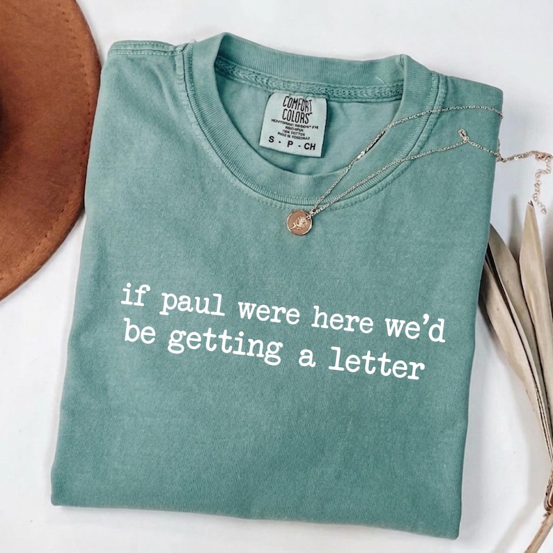 Może przedstawiać: Miętowa koszulka z białym napisem "if paul were here we'd be getting a letter". Złoty naszyjnik z wisiorkiem spoczywa na koszulce. Koszulka marki Comfort Colors, rozmiar S.