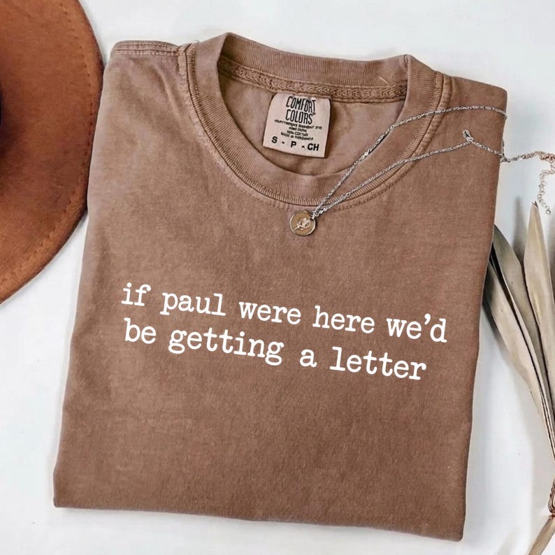 Może przedstawiać: Brązowa koszulka Comfort Colors z białym napisem "if paul were here we'd be getting a letter". Srebrny naszyjnik z małą zawieszką leży na koszulce. Koszulka jest złożona.