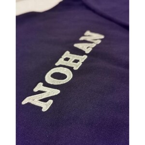 Peut inclure: Gros plan sur un vêtement bleu foncé avec le mot "NOHAN" brodé en blanc. Le texte est en gras, en police sans-serif, et le tissu a une texture subtile. L'image est bien éclairée, soulignant les détails de la broderie.