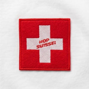 Peut inclure: Un écusson carré rouge avec une croix blanche centré sur un fond en tissu blanc. La croix blanche présente le texte brodé en rouge HOP SUISSE! au centre. L'écusson a une bordure cousue visible et un aspect texturé semblable à du feutre.