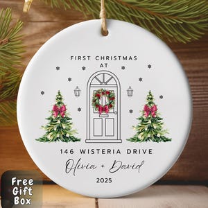 May include: A white ceramic Christmas ornament with a festive design. It features a door with a wreath, Christmas trees, and the text "First Christmas At" and "146 Wisteria Drive Olivia + David 2025".
