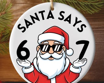 May include: A round Christmas ornament with the text "SANTA SAYS" and the numbers "6 7". The ornament features a cartoon Santa Claus wearing sunglasses and a red suit. The ornament is hanging from a string.