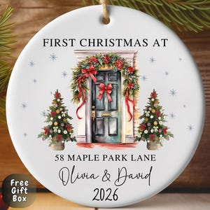 May include: A round white ceramic Christmas ornament featuring a printed illustration of a front door decorated with a festive wreath and two small Christmas trees. The text reads FIRST CHRISTMAS AT 58 MAPLE PARK LANE Olivia & David 2026.
