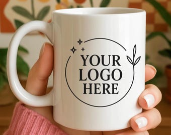 May include: White ceramic mug with a curved handle. The mug features a black circular logo design with the text "YOUR LOGO HERE", along with stars and a leaf. The background includes floral art and potted plants.
