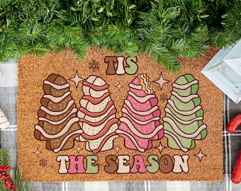 May include: A rectangular doormat with a festive design. The mat features four stylized Christmas trees in shades of brown, pink, and green, with the text "TIS THE SEASON". The mat is surrounded by holiday decorations, including candy canes and greenery.
