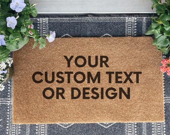 May include: A rectangular brown coir doormat with the text "YOUR CUSTOM TEXT OR DESIGN" in black. The mat is placed on a patterned gray rug, with potted flowers on either side.