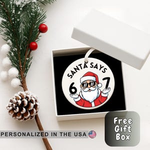 May include: A white ceramic Christmas bauble with a Santa Claus design. The bauble features Santa wearing sunglasses and a red suit, with the text "SANTA SAYS 6 7". The bauble is in a white gift box.