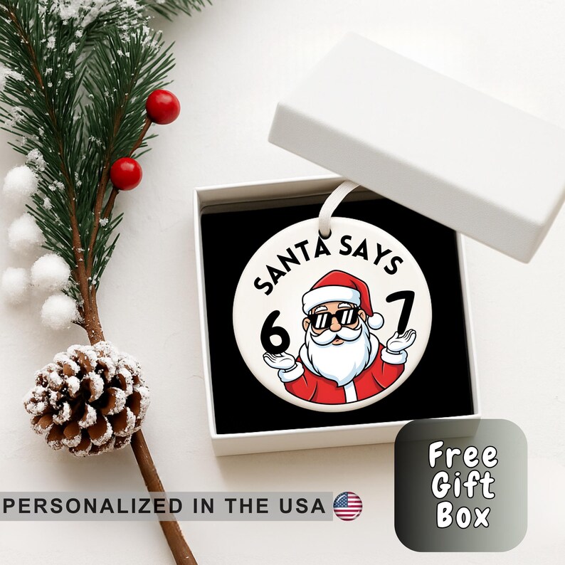 May include: A white ceramic Christmas bauble with a Santa Claus design. The bauble features Santa wearing sunglasses and a red suit, with the text "SANTA SAYS 6 7". The bauble is in a white gift box.