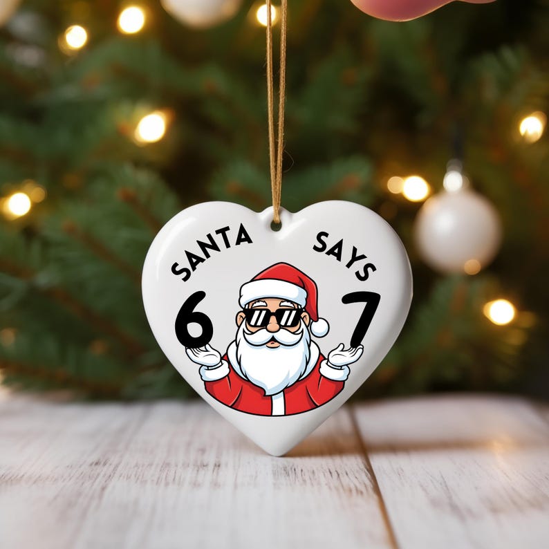 May include: White heart-shaped Christmas ornament with the text "SANTA SAYS 6 7". The ornament features a cartoon Santa Claus wearing sunglasses. It has a brown string for hanging, and is suitable for a Christmas tree.