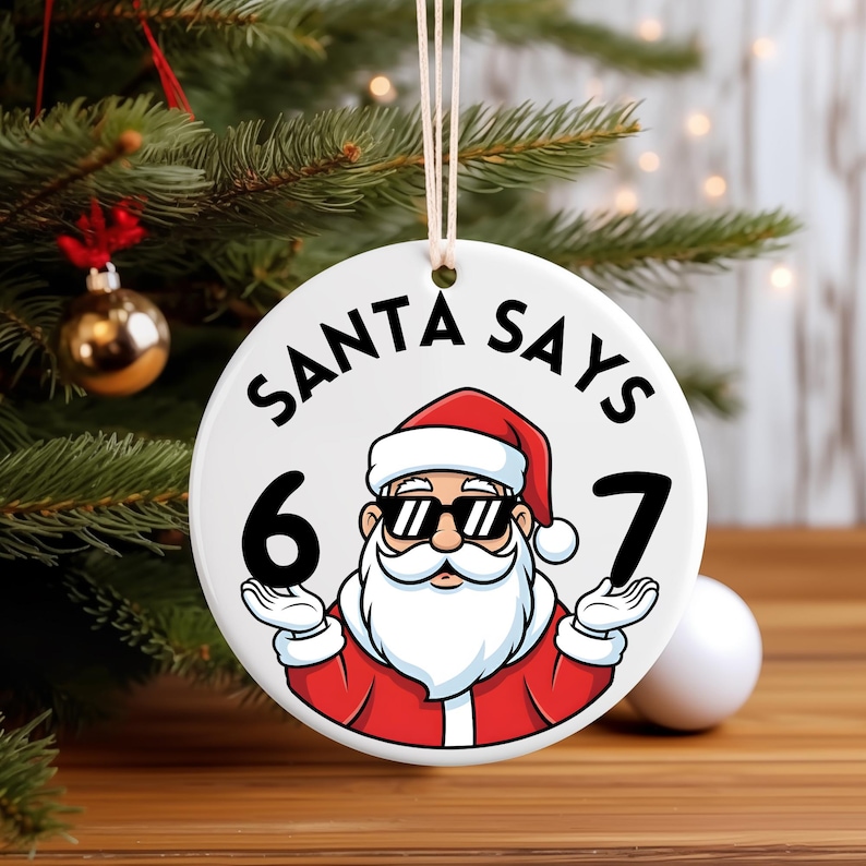 May include: A circular Christmas tree decoration featuring a cartoon Santa Claus wearing sunglasses and holding up the numbers 6 and 7. The text "SANTA SAYS" is above the Santa image. The ornament is white with a hanging string.