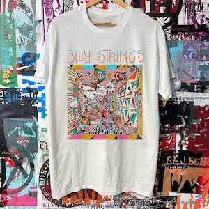 May include: White t-shirt with the text "BILLY STRINGS" in colorful letters. The shirt features a vibrant, psychedelic graphic design with various shapes and colors, including pink, orange, and blue. The design is centered on the front of the shirt.