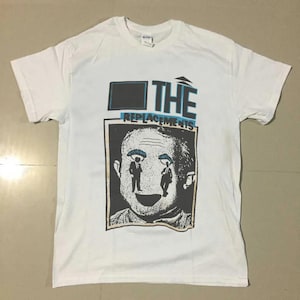 May include: White t-shirt featuring a graphic of a face with two figures in the eyes and the band name "THE REPLACEMENTS" in blue and black. The shirt is a short-sleeved crew neck.