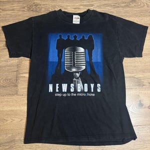 Może przedstawiać: Czarna koszulka z grafiką mikrofonu, sylwetkami zespołu oraz napisem "NEWSBOYS" i "step up to the microphone". Projekt jest w kolorze niebieskim i białym. Koszulka ma okrągły dekolt i krótkie rękawy.