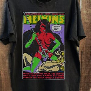 Op de afbeelding: Zwart t-shirt met een afbeelding van een rode duivelachtige figuur met een drietand. De afbeelding bevat de tekst "MASTERS OF ROCK MELVINS" en "INVERTED CROSSES MARK THE GRAVES".
