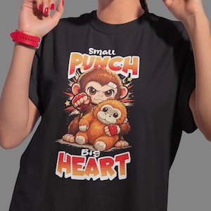 Peut inclure: T-shirt noir avec une illustration de dessin animé de deux singes. Le singe du haut est brun avec un bandage et un poing serré. Le singe du bas est orange. Le texte dit "Small Punch Big Heart".
