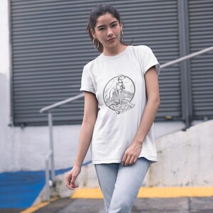 Può includere: T-shirt bianca con un'illustrazione in bianco e nero di un cane su una montagna, incorniciata da un cerchio. Il cane indossa un collare e si trova su una superficie a motivi. La maglietta ha maniche corte e girocollo.