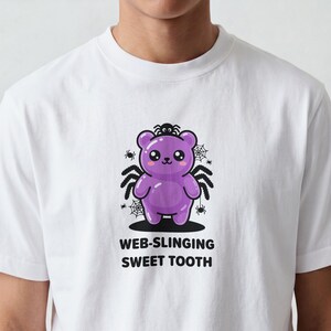 Peut inclure: Un t-shirt blanc avec un motif représentant un ours en gélatine violet avec des pattes d'araignée noires, de petites araignées et des toiles d'araignée. Le texte sous le motif indique WEB-SLINGING SWEET TOOTH en lettres capitales noires.