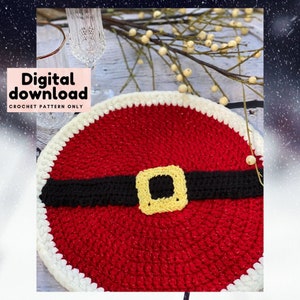 May include: A red and white crochet coaster with a black belt and a yellow buckle, resembling Santa Claus's belt. The coaster is on a wooden surface with a white branch with small white berries.