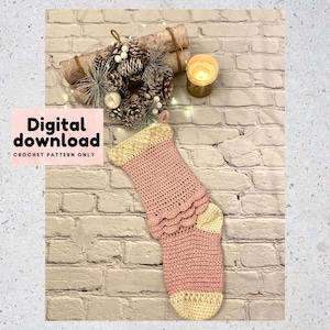 Op de afbeelding: Een roze gehaakte kerstsok met een witte gehaakte boord en een gegolfde rand. De sok hangt aan een bakstenen muur met een dennenappelenkrans en een kaars op de achtergrond. De tekst "Digitale download HAKELPATROON ALLEEN" staat op de afbeelding.